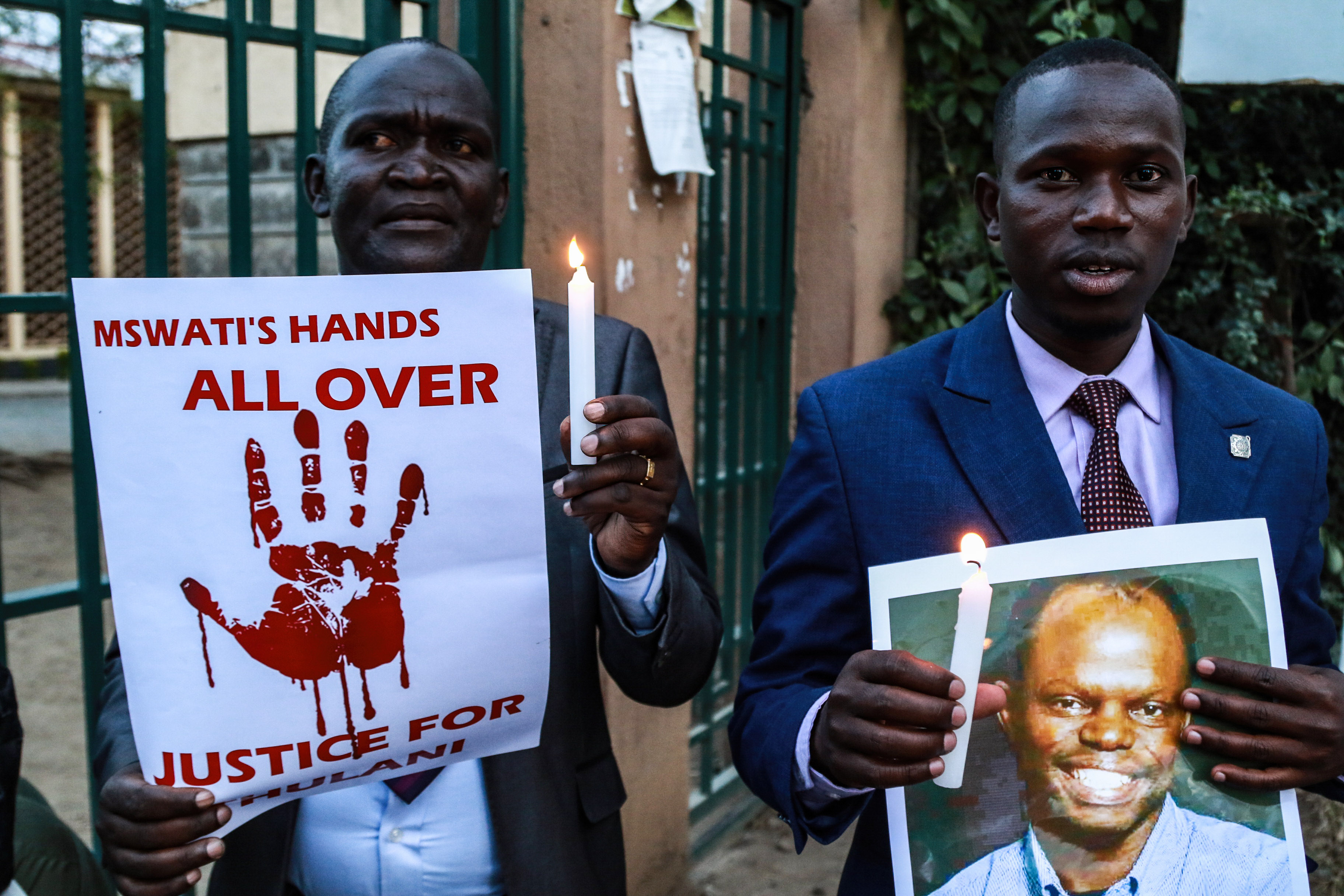 Two men stand in front of a gate holding candles and posters. One poster has an image of a bloody handprint and reads, "Mswati's hands all over, Justice for Thulani." The other poster is a photo of a smiling man.