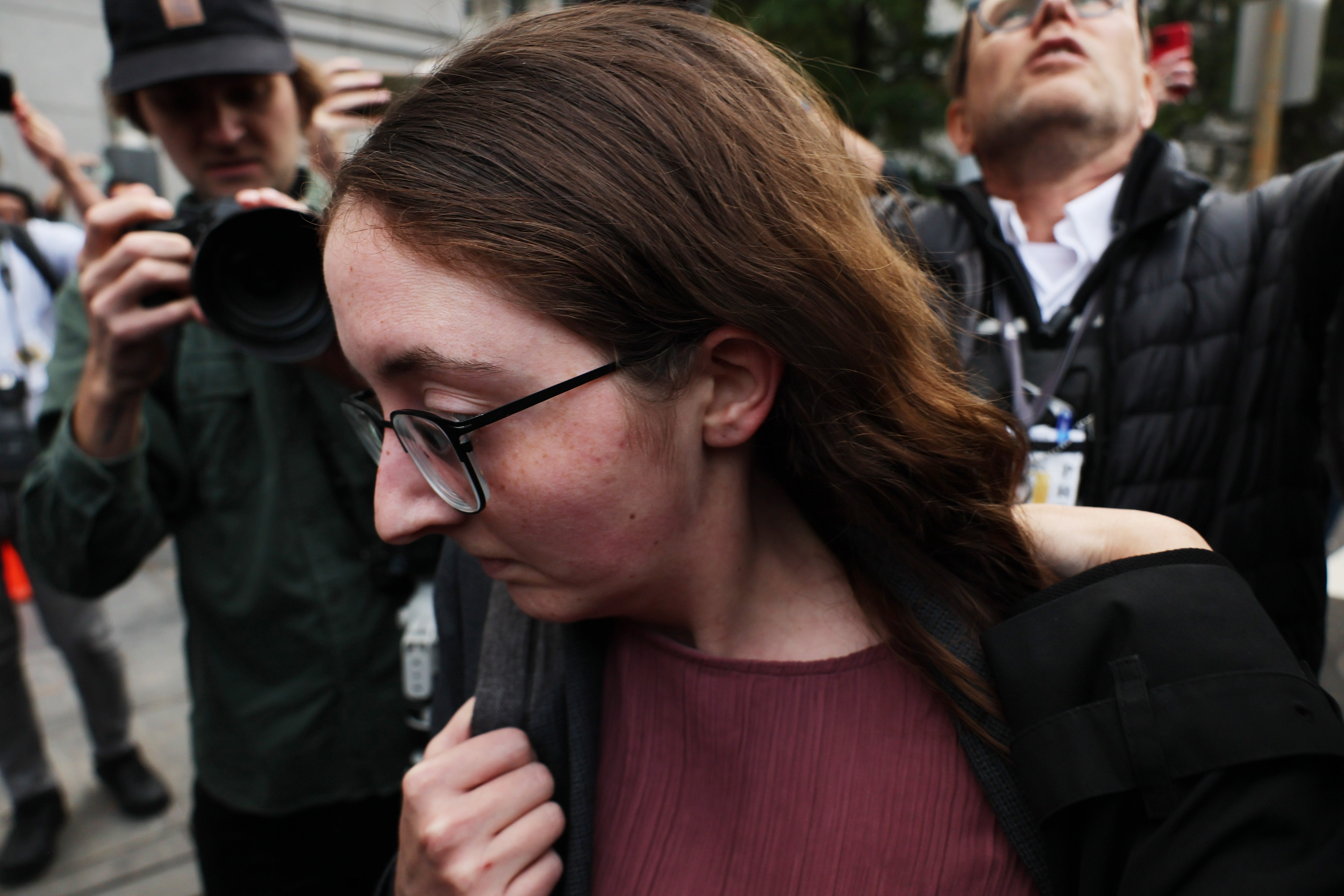A young woman with flat long brown hair and glasses looks down as she walks away from media with cameras.