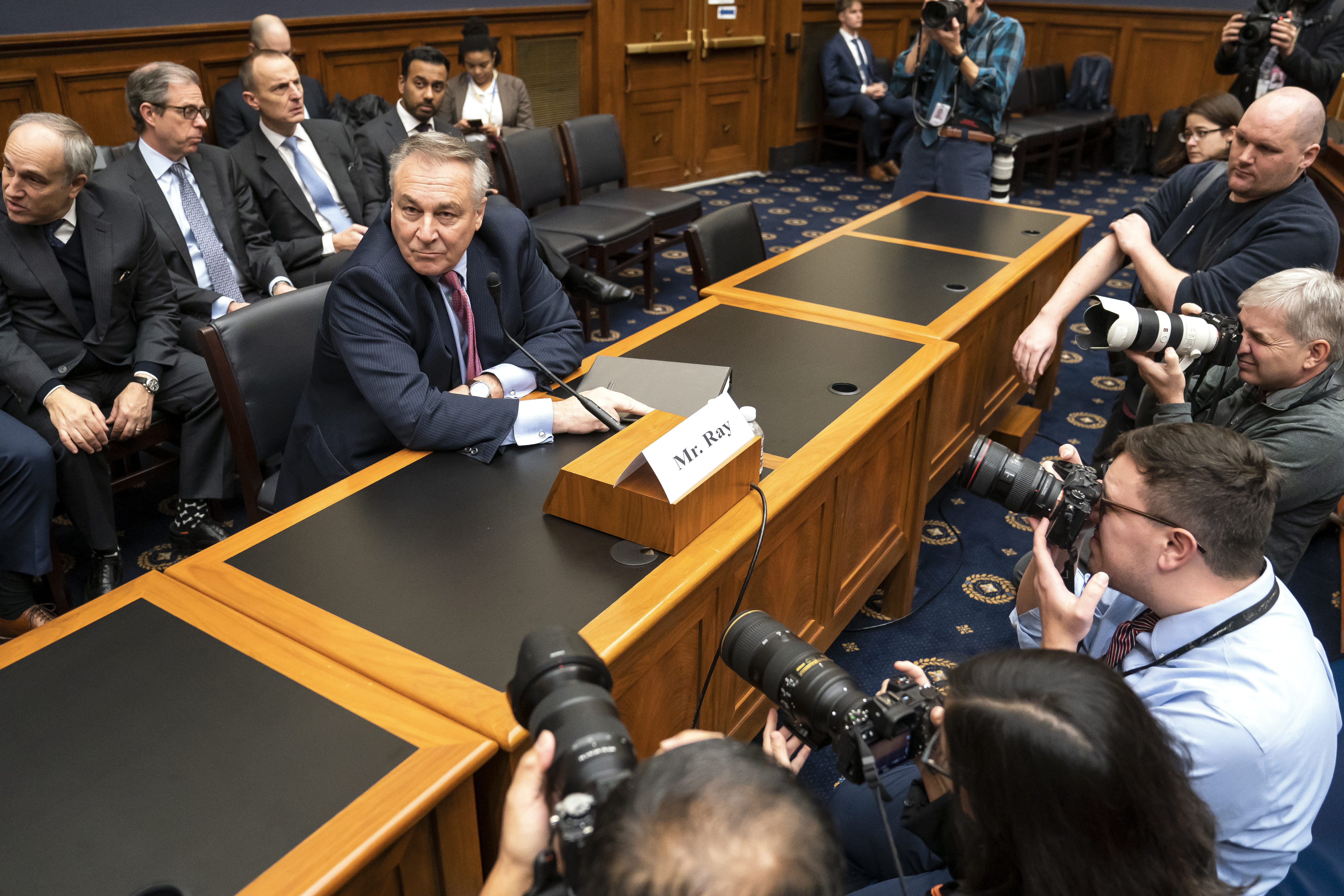 A gray-haired man in a blue business suit and purple tie sits behind a table as photographers kneel in front of him on the other side. Similarly aged men sit behind him in several rows of chairs.