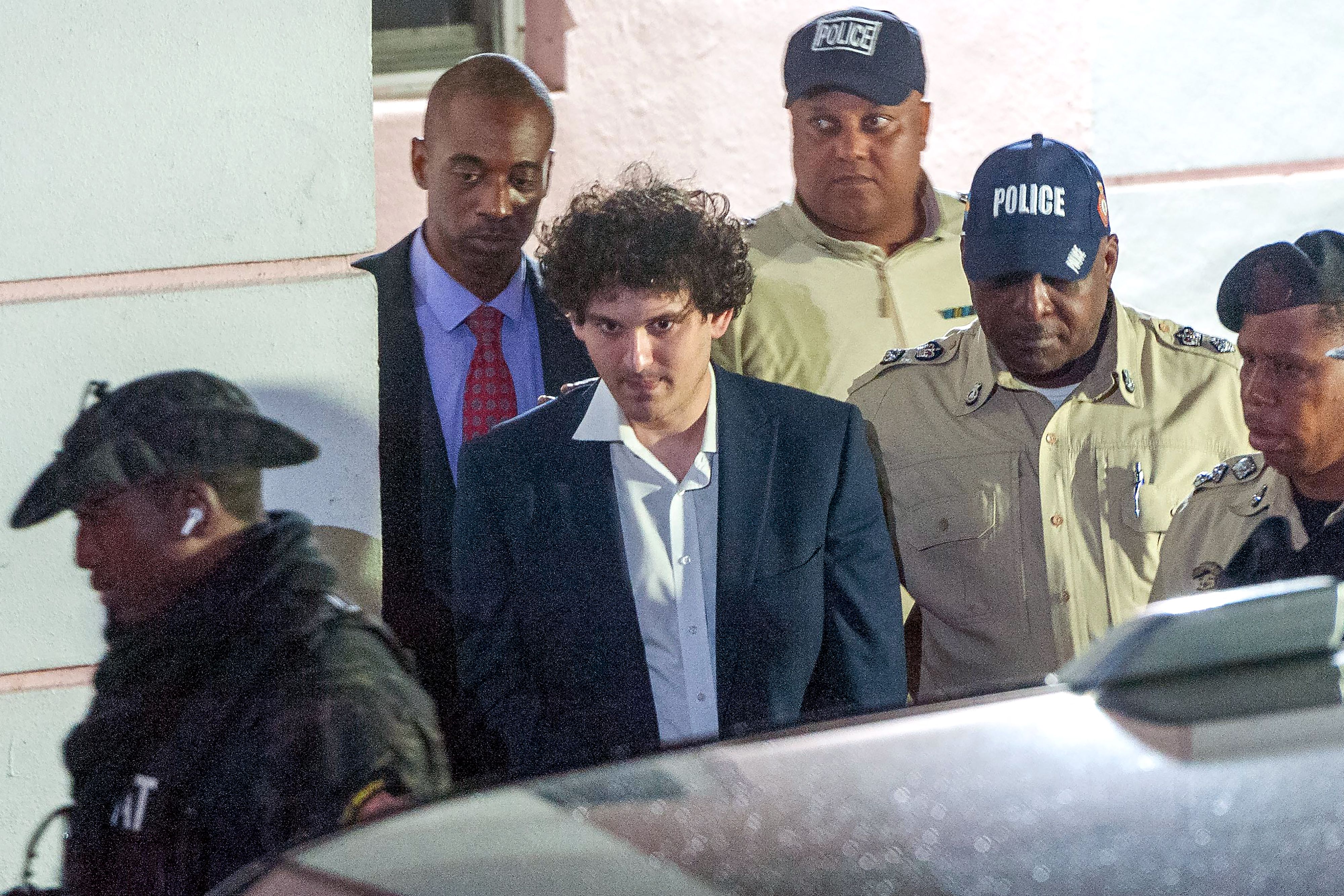 A man with thick curly hair in a suit jacket and a shirt with splayed collars stares blankly as he's escorted by police officers.