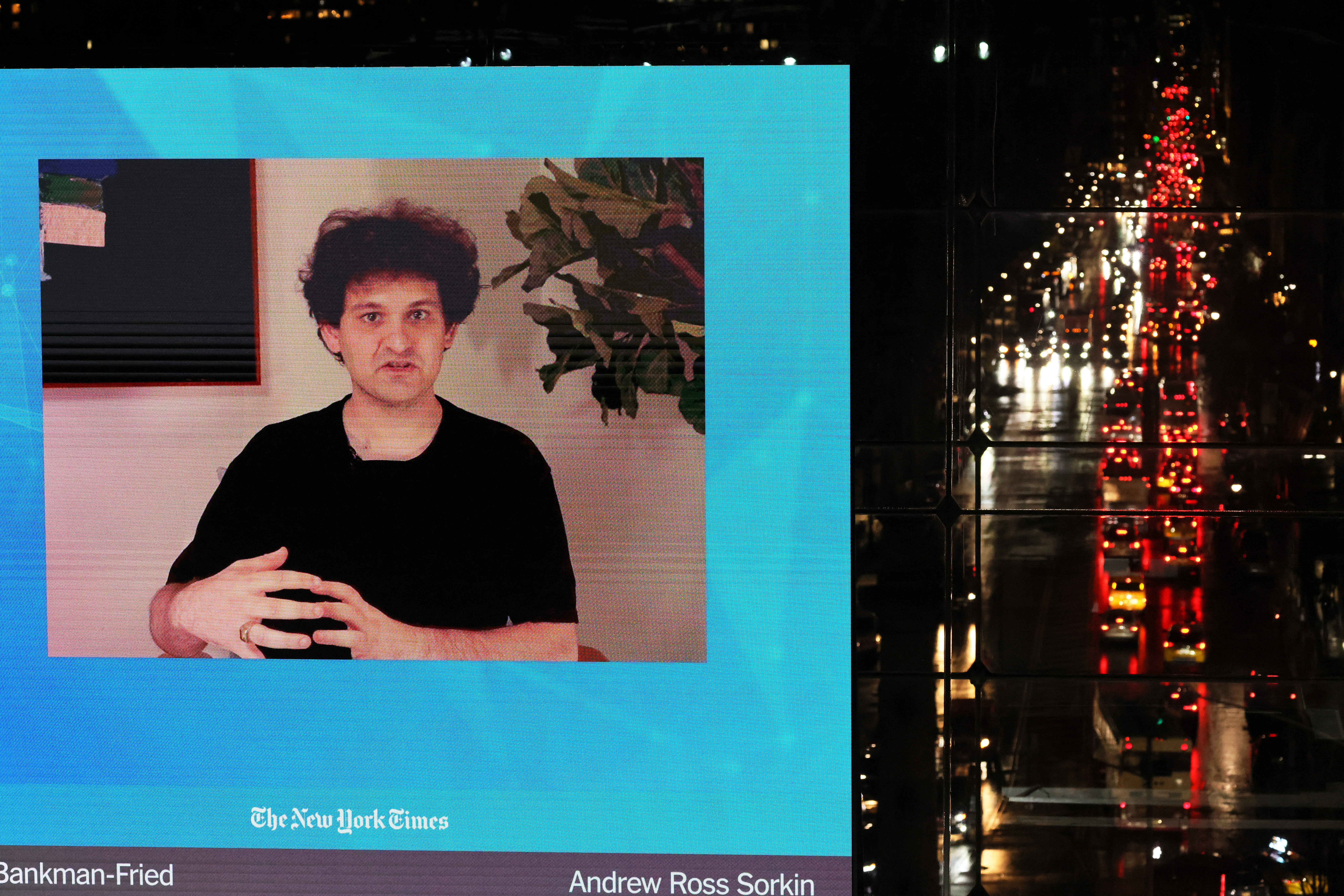 A young man with thick curly hair and black t-shirt is seen speaking on a screen. On the screen are the words "The New York Times," "Bankman-Fried" and "Andrew Ross Sorkin."
