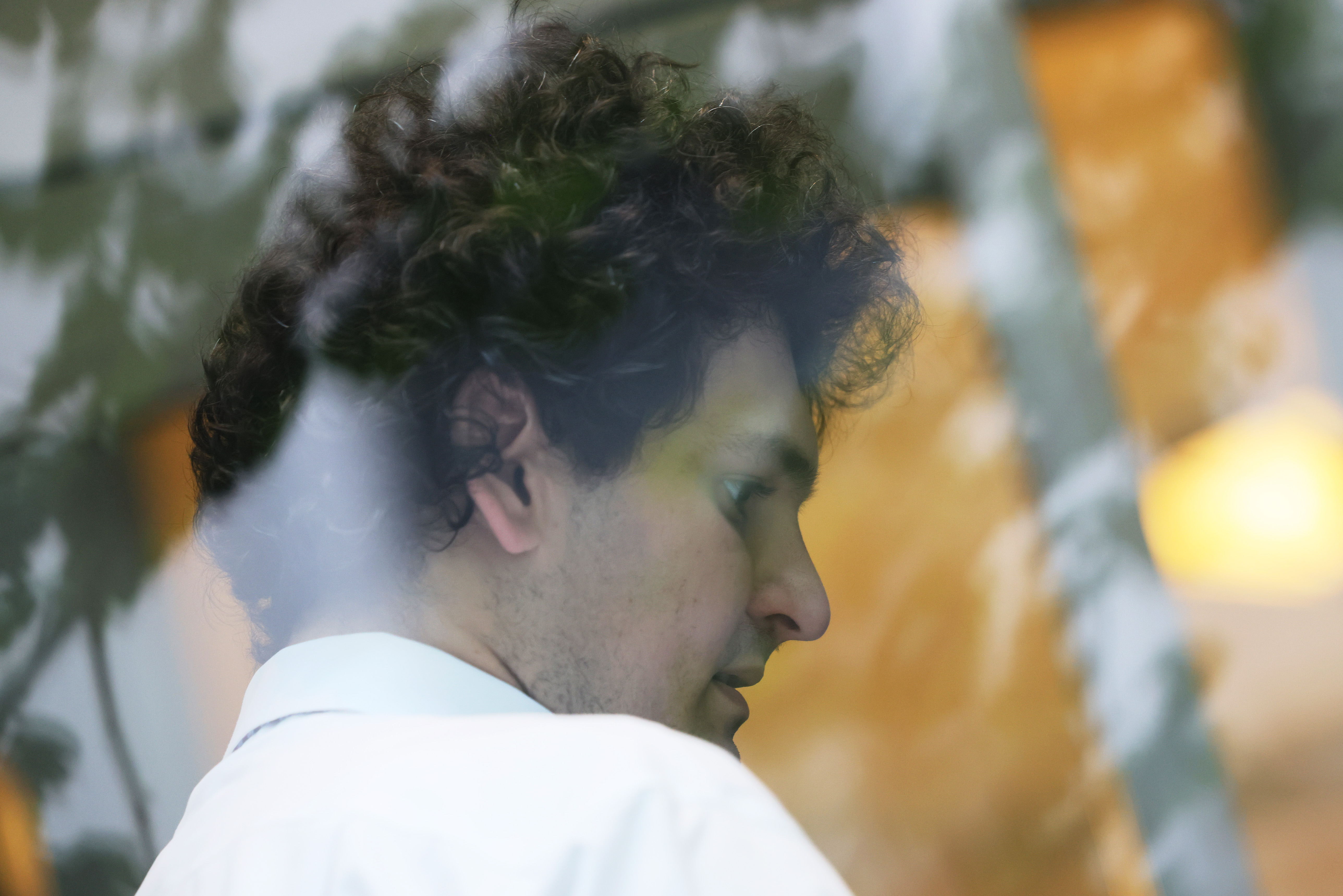 A man with thick curly hair, seen wearing a white shirt, looks downward. The man stands on the other side of glass window, which reflects leaves on a tree.
