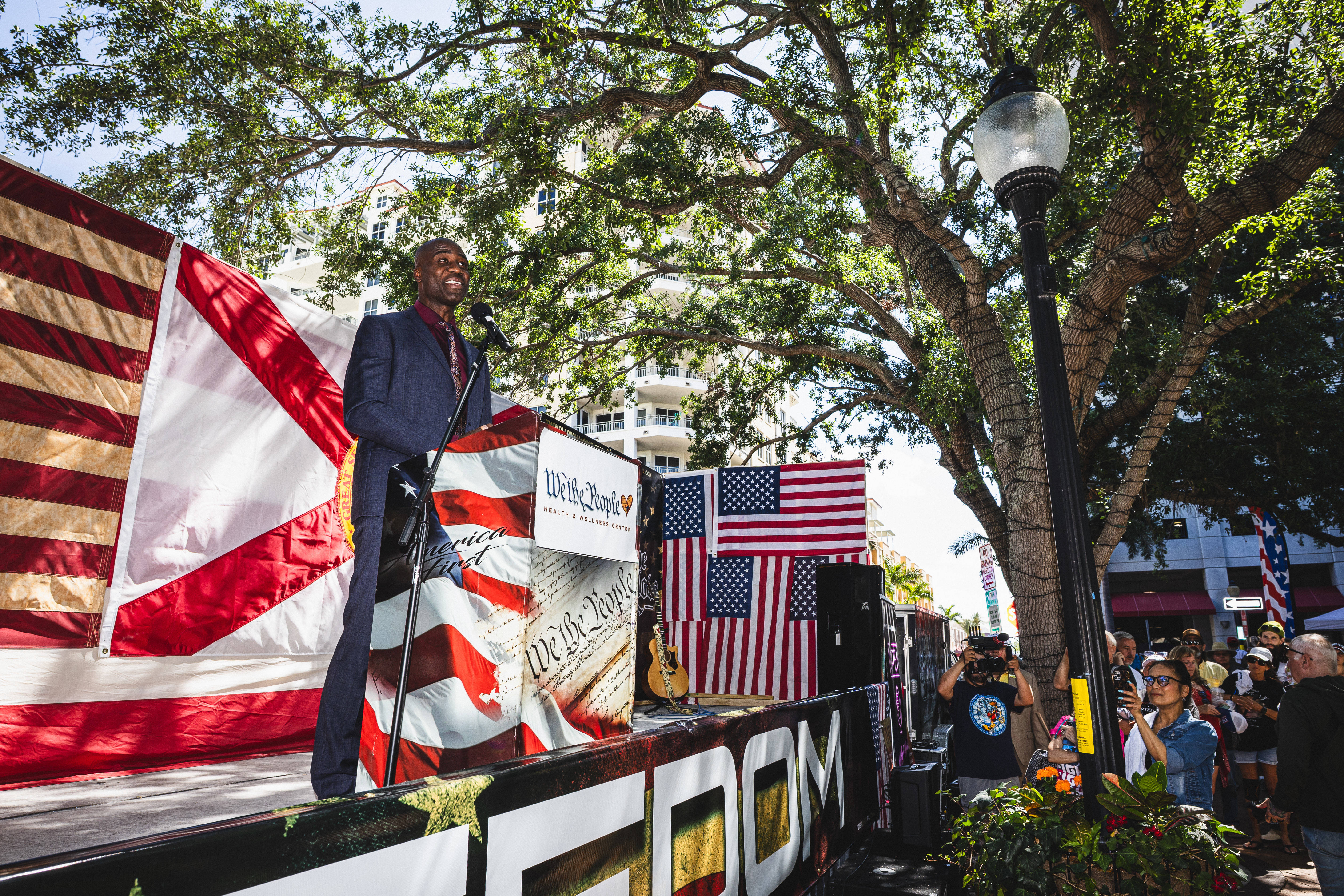 A black man in a suit stands on a stage outdoors underneath a tree. The stage is layered with American flags, the Florida flag and signs that read "We the People."