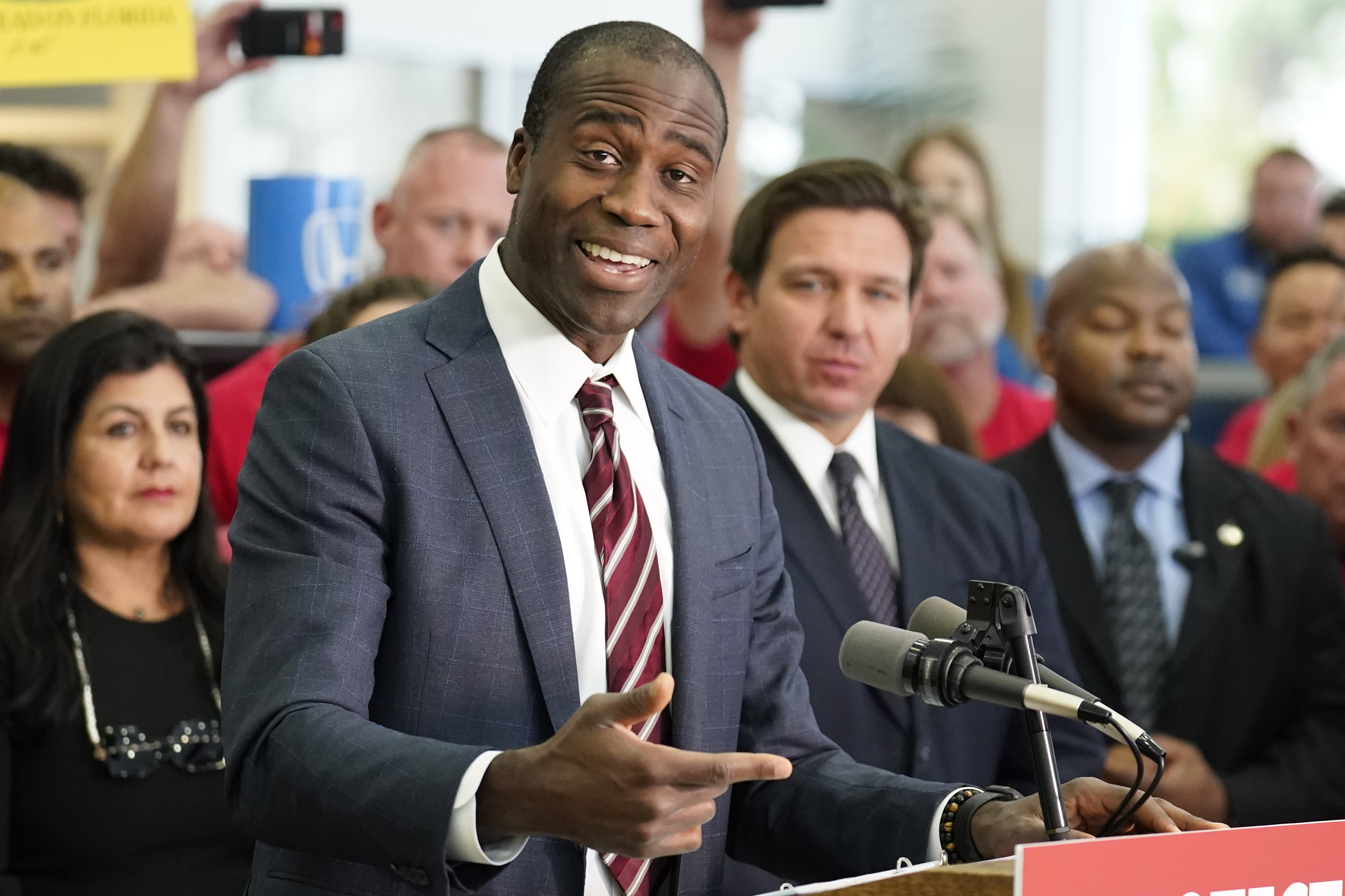 A Black man in a suit speaks as he stands behind a lectern. A crowd of people stand behind him, listening intently, some with smiles on their face.