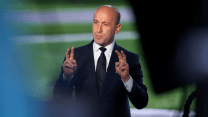 A bald man in a suit makes air quotes with his fingers while speaking.