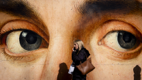 A Caucasian woman with hair down to her shoulders, wearing a coat and carrying two bags, walks to our left as she talks on a mobile phone. Behind her is a large mural of a face, seen from the bridge of the nose to the forehead. The dark gray eyes of the painting seem to be in line with the passing woman, whose shadow falls against the lower part of the wall.