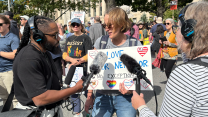 A man with long dreadlock hair and bushy beard holds a microphone in front of a woman with short brown hair. She is among a crowd of people wearing sunglasses, T-shirts, jeans and shorts, who are holding signs. The woman’s sign reads, “Love Your Neighbor. No Exceptions.”