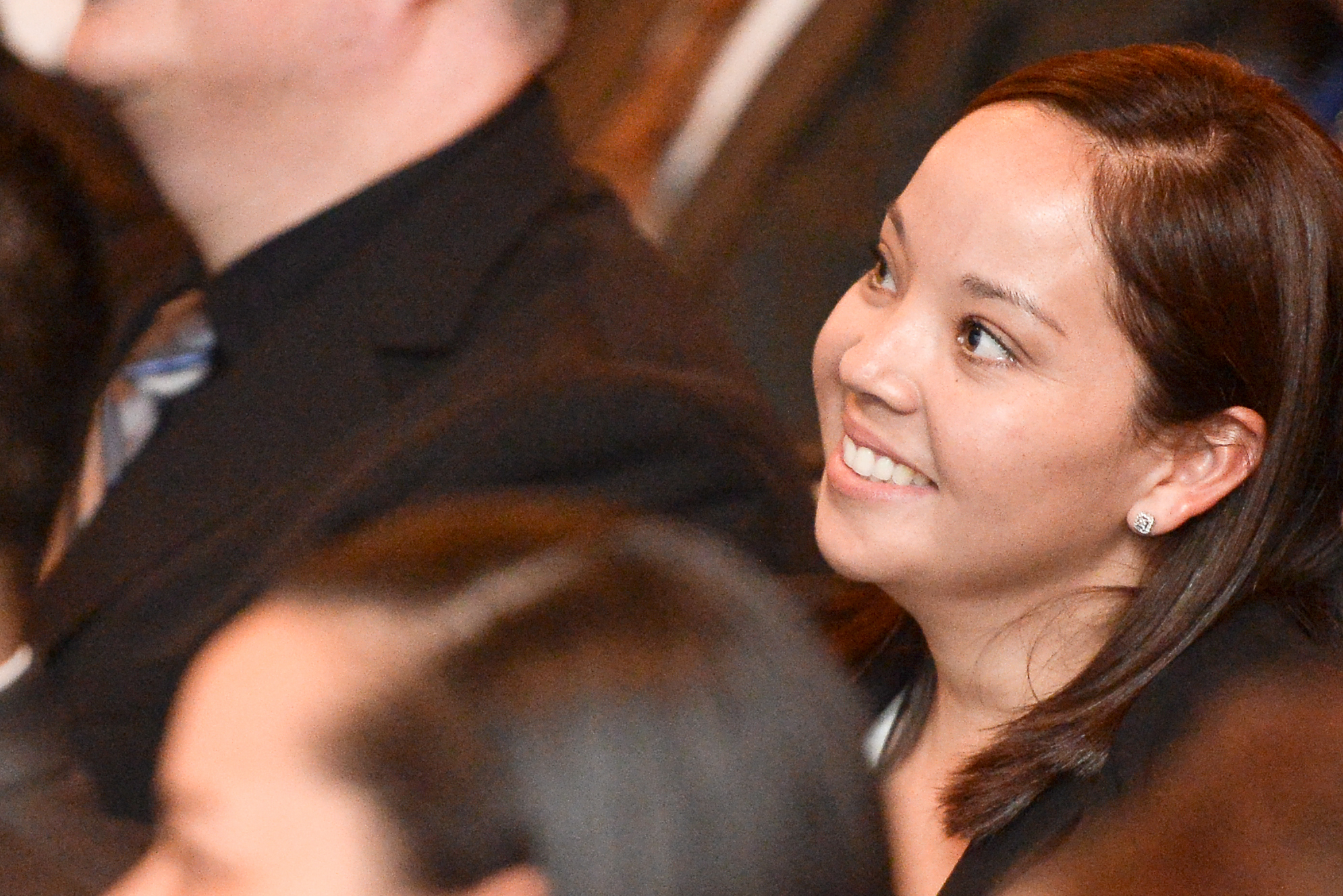 The face of a young smiling woman with dark brown hair is seen in a crowd of other people