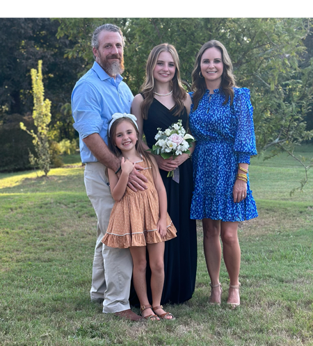 A man in long sleeve blue shirt and khakis poses with his wife, teenage daughter and young daughter, who all wear dresses. They pose outside, the teenage daughter holds a bouquet of flowers.