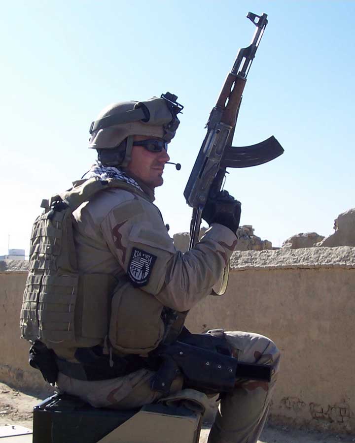 A soldier in a brown desert camouflage military uniform outdoors holds a gun.
