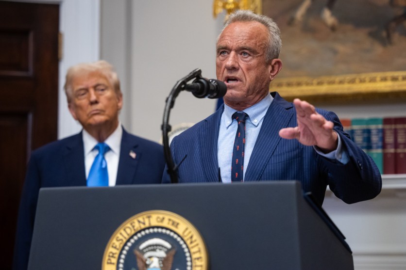 President Donald Trump and Robert F. Kennedy Jr. speak a the White house. Kennedy is a lectern motioning with his left hand and Trump standing off to the side, making that face he makes where you can tell he's probably thinking about something else entirely.