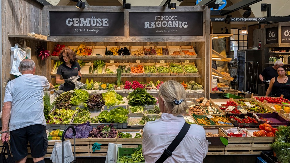 Two people stand in front of a market stall of vegetables.