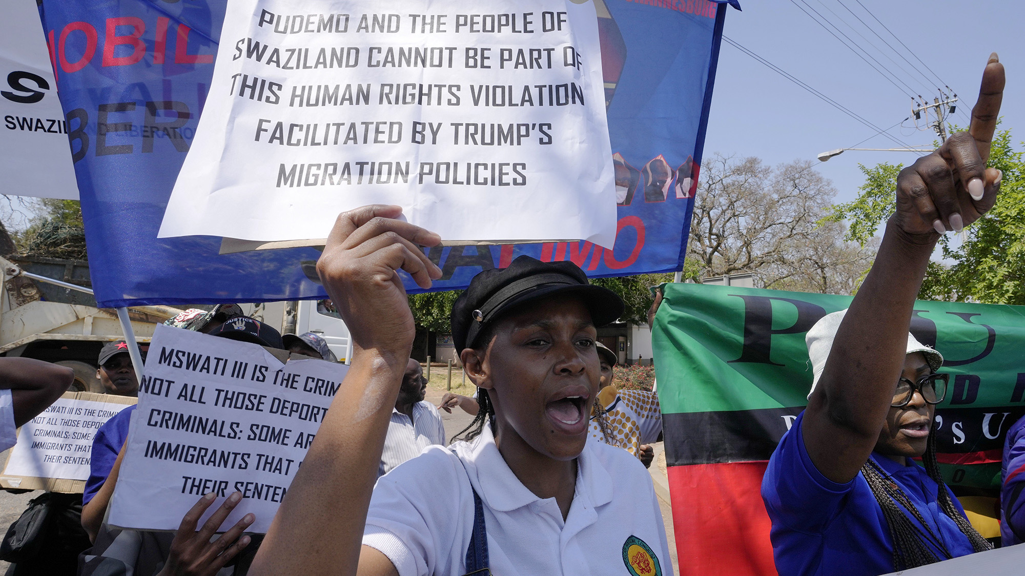 A group of people hold various signs and banners. One sign in the foreground reads, "Pudemo and the people of Swaziland cannot be part of this human rights violation facilitated by Trump's migration policies."