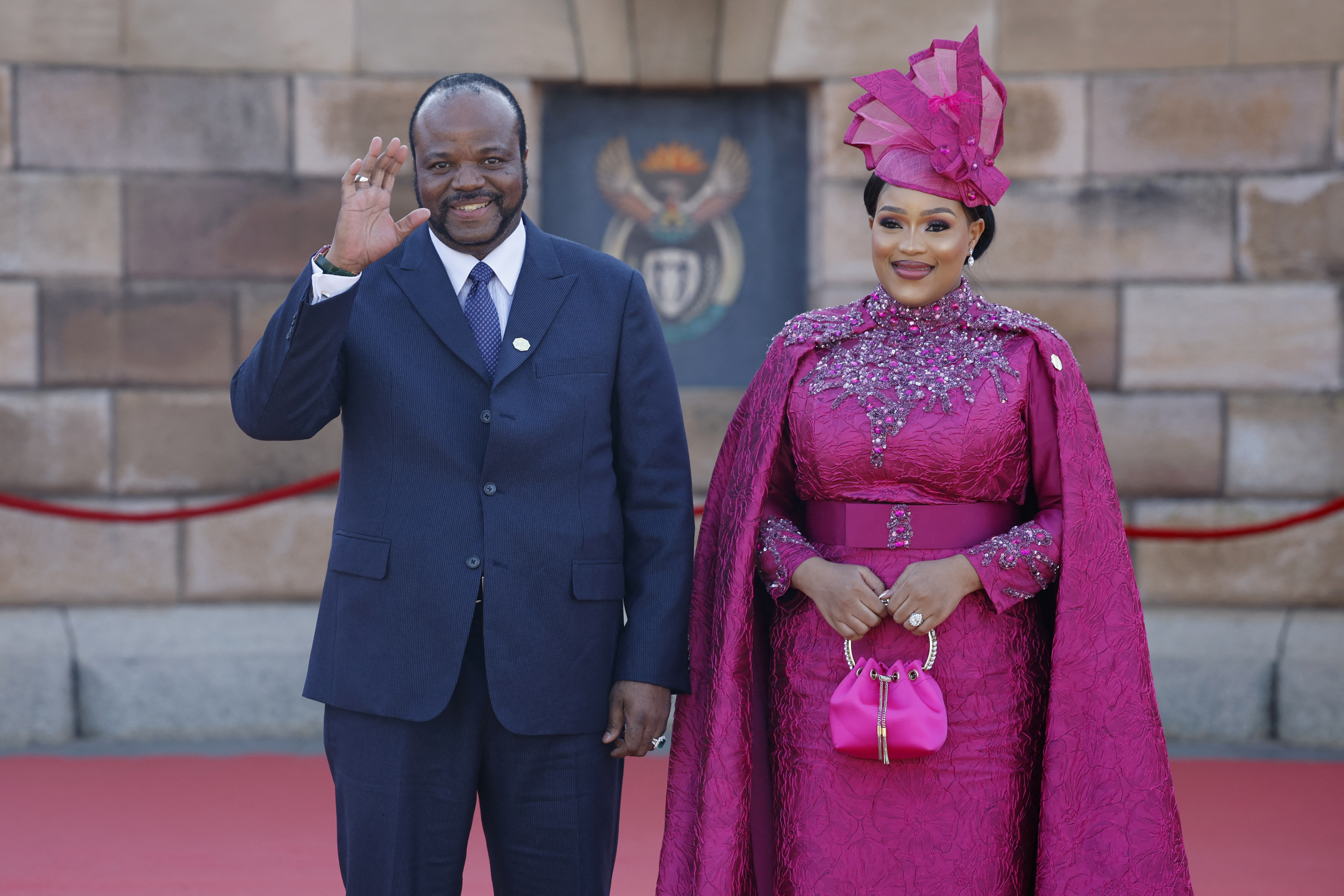 Smiling man in a dark blue suit, waving, standing beside a smiling woman dressed in a formal, bejeweled pink gown with a matching cape, handbag, and hat.