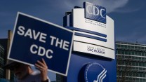 A person holds a "Save the CDC" poster in front of a large blue and white CDC sign on CDC campus