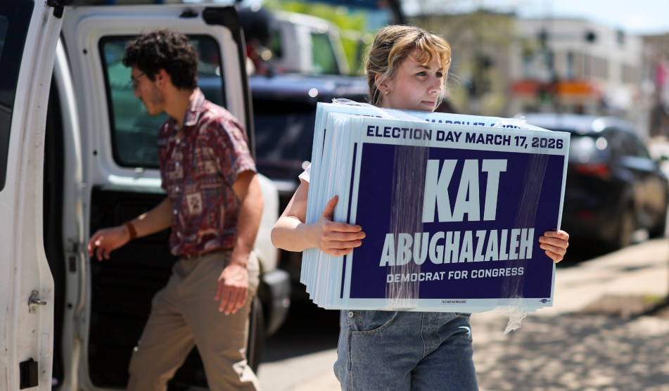 a woman holding a sign in front of a Kat for Congress sign on a street