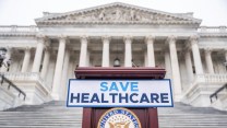 A sign on a podium that says "Save Healthcare" in front of an old building.