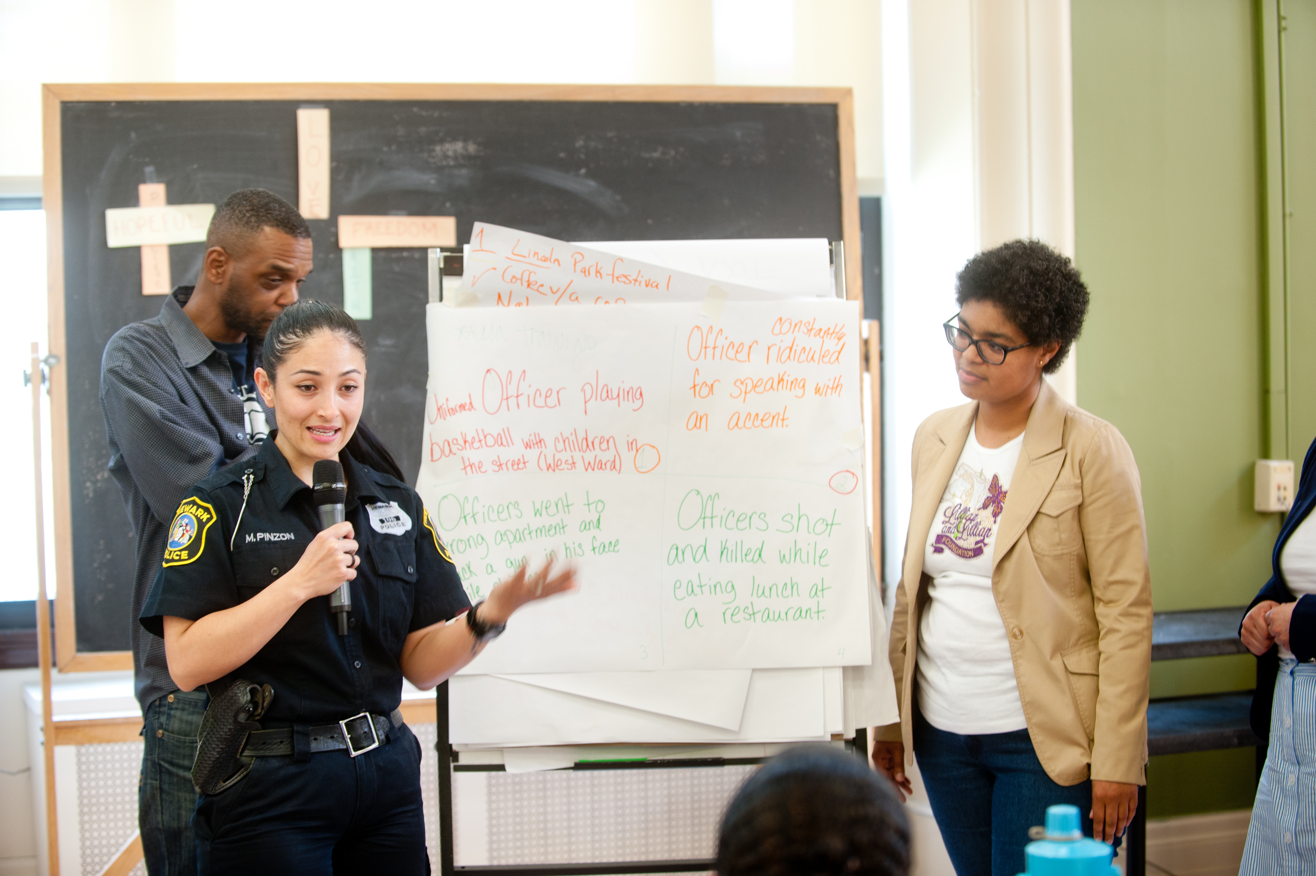 A female police officer speaks to community members at a EJUSA event in front of a whiteboard outlining key topics.