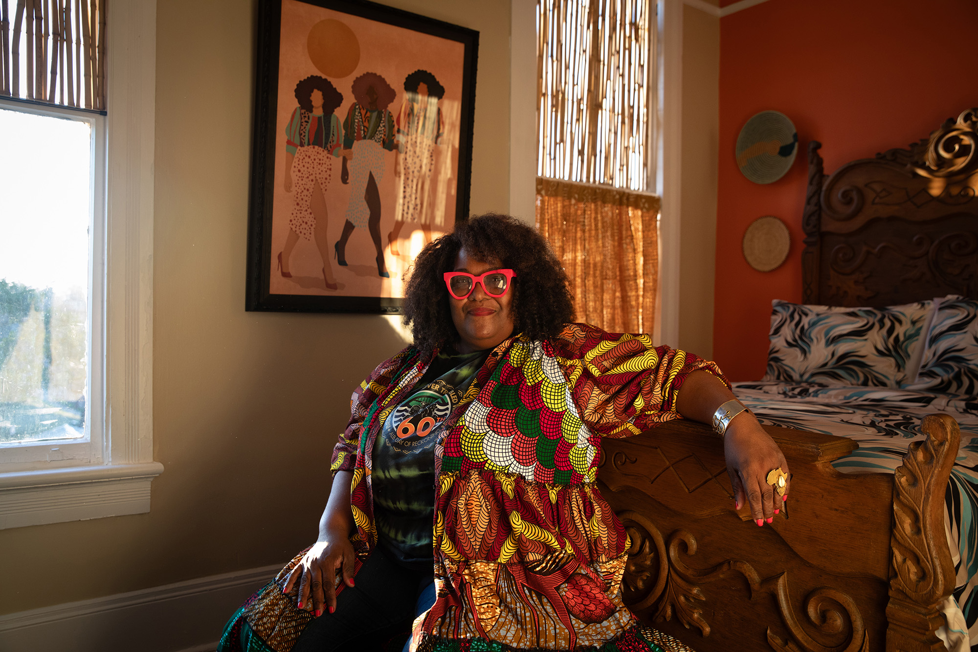 Portrait of African American woman wearing red glasses, sitting with her arm drapped over a bed.