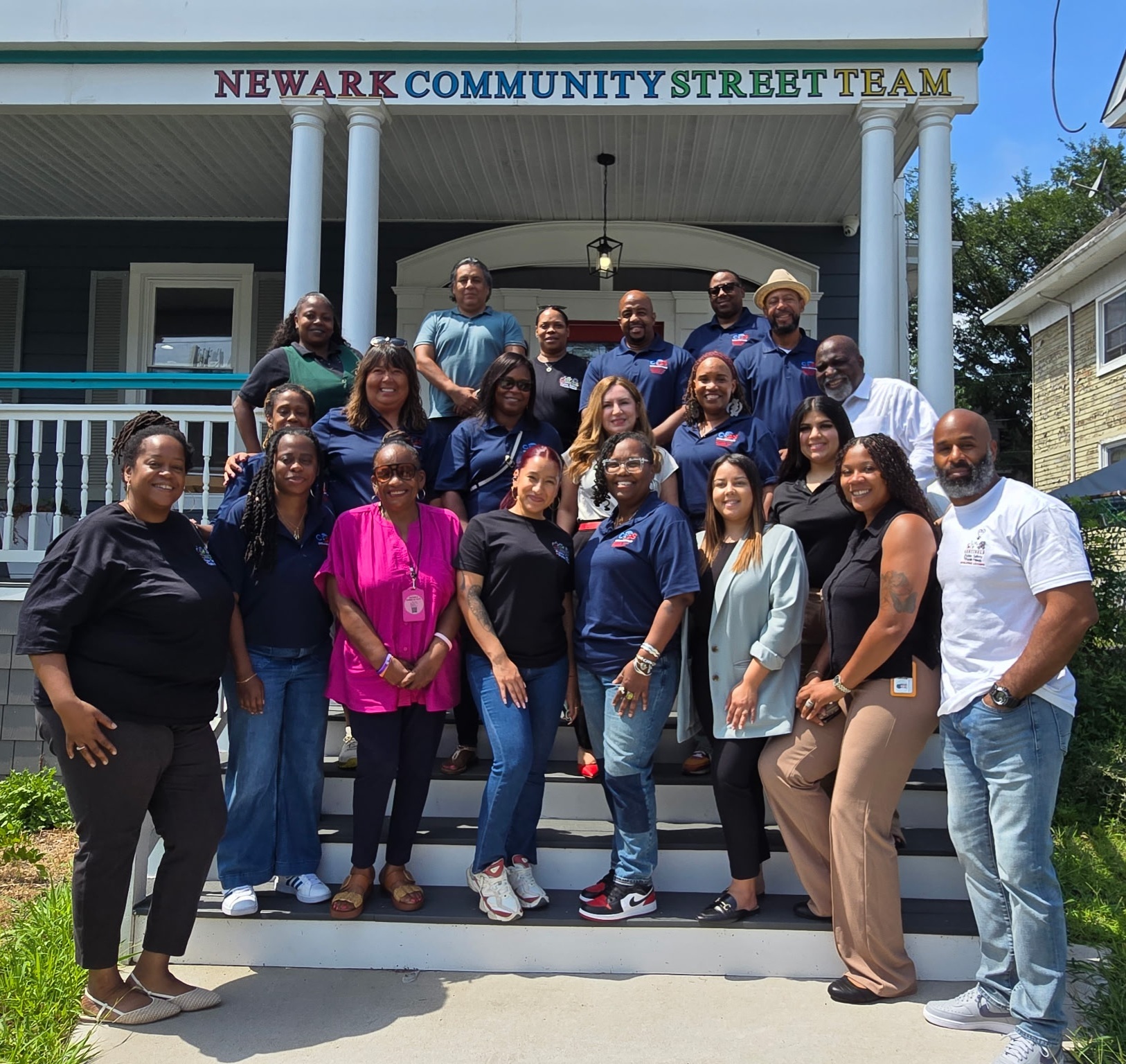 Group of staff members of varying age and diversity stand smiling on the stairs of the Newark Community Street Team building.
