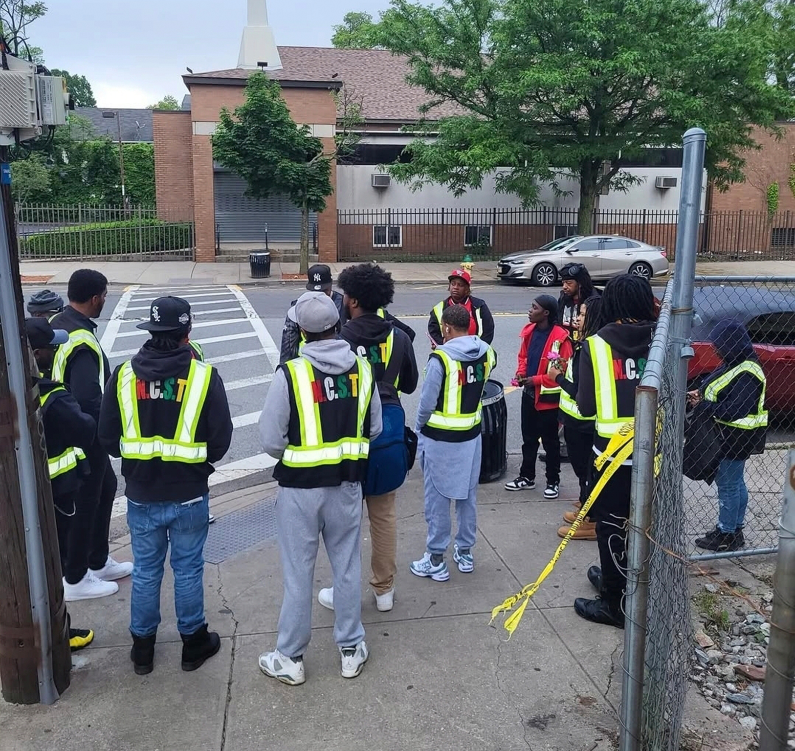 A group of diverse Newark Community Street Team workers wearing high-visibility vests gather to discuss their tasks on the sidewalk.