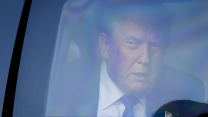 A close-up photograph of Donald Trump peering out a back window of the presidential limousine. With narrowed eyes and mouth slightly ajar, he is wearing a suit and tie with an American flag pin on his left lapel.