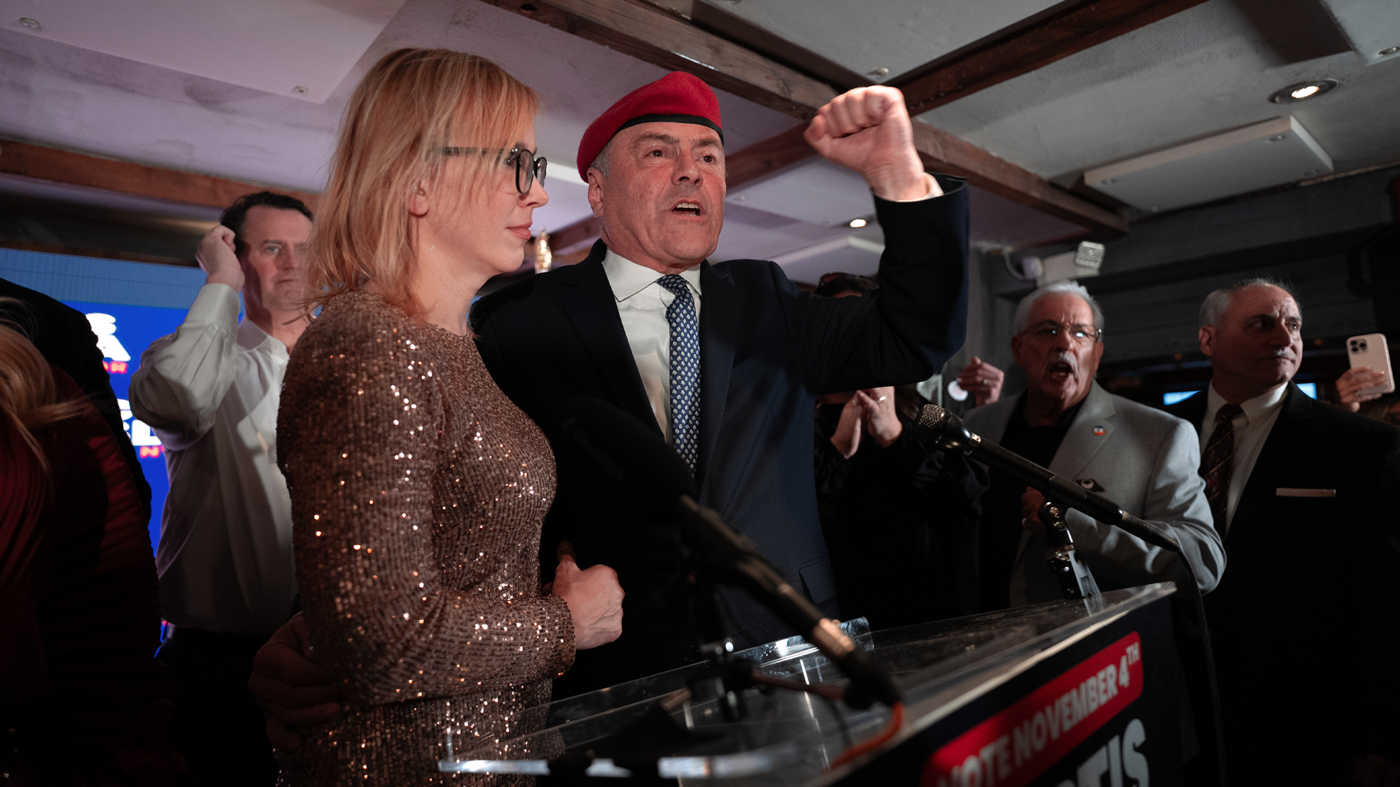 A Caucasian man in a suit and red beret pumps his fist as he stands at a lecturn with supporters gathered around him. Most of the supporters are men in suits. His wife has shoulder-length brown hair and stands beside him in a bronze, glittery dress.