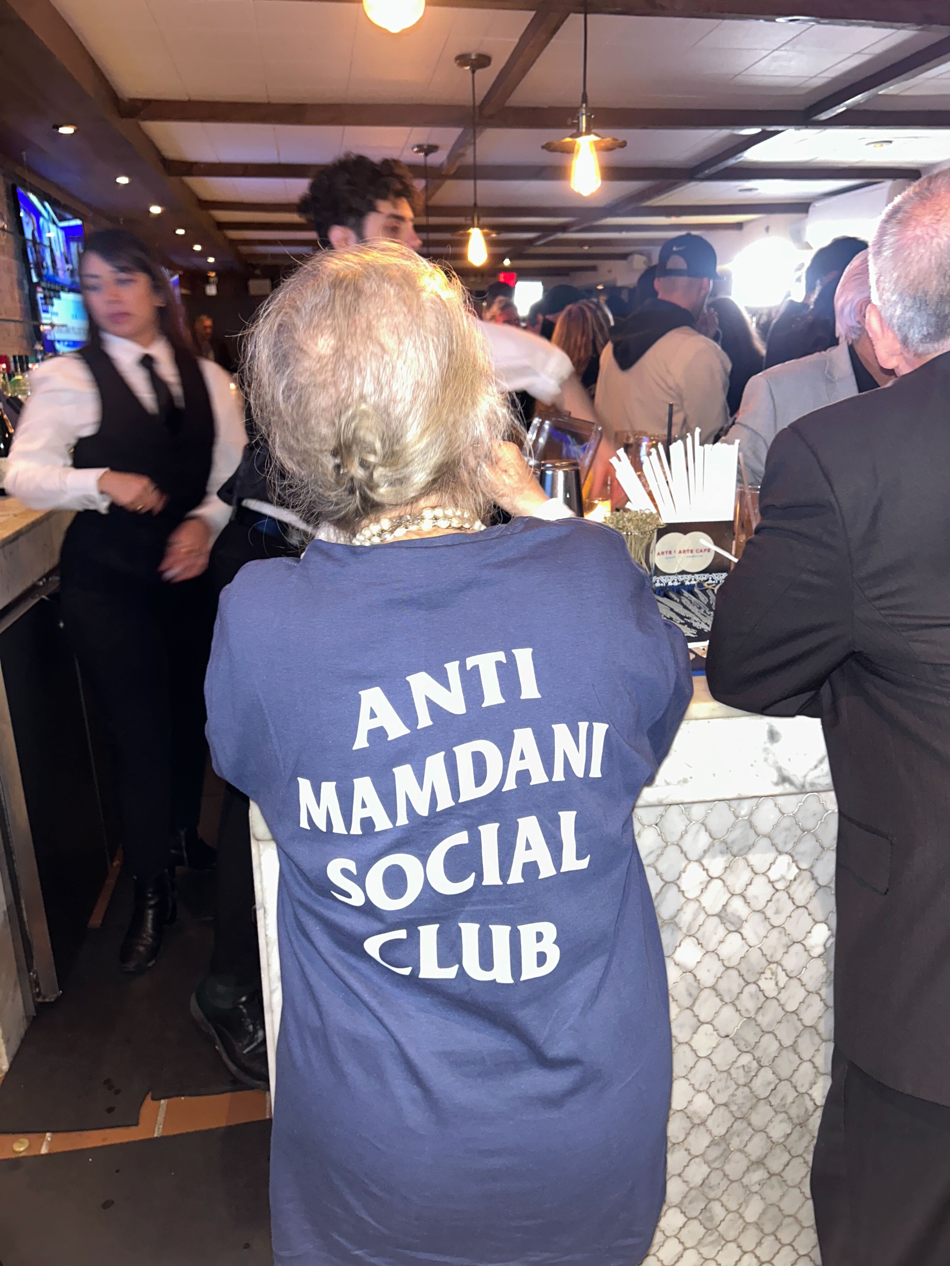 An elderly woman with her hair in a bun is seen from behind wearing a blue t-shirt with 'Anti Mamdani Social Club' written in white serif letters on the back. The woman is standing at a crowded bar where people are being served.