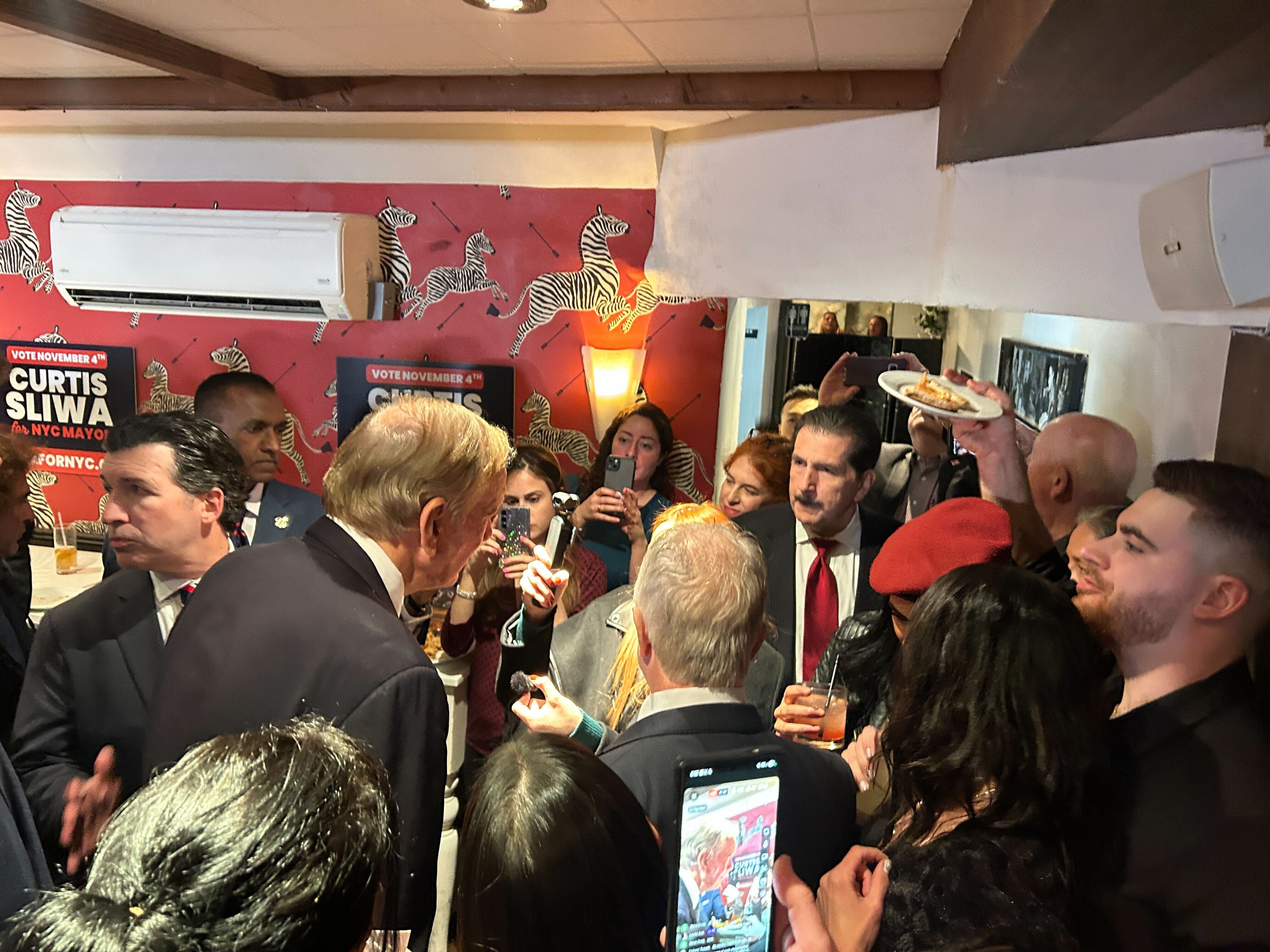 Women, along with men in suits, crowd around an elderly Caucasian male in a room that has red, white and blue campaign signs in support of Curtis Sliwa. Many people have their mobile phones out to record the moment. The man at the center, George Pataki, leans in to talk into a small microphone a woman holds in front of him.