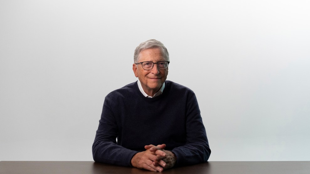 A white man sits at a table in front of a white background and smiles faintly.