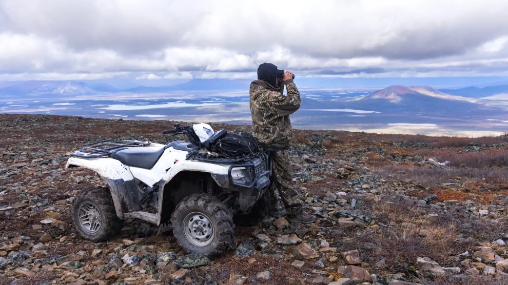 A person next to a white four wheeler looks at the landscape through binoculars.