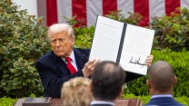 Trump sits at a desk outside the White House, holding up a two page document he just signed.