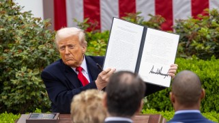 Trump sits at a desk outside the White House, holding up a two page document he just signed.