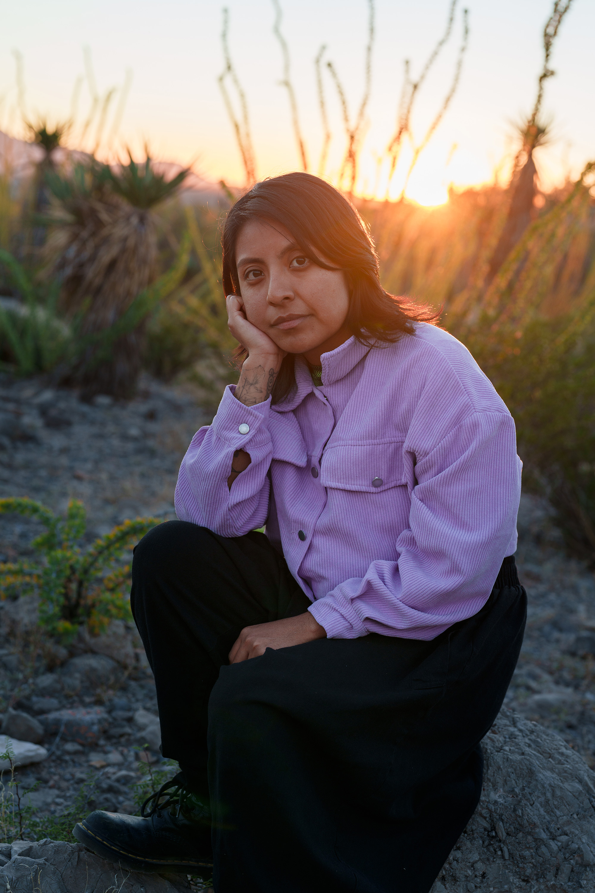 Woman kneeling for a portrait in the desert at sunset. She has shoulder-length dark hair and wears a lilac-colored jacket and dark pants.